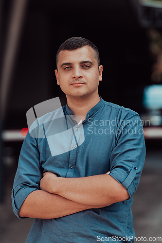 Image of A successful young businessman in a shirt, with crossed arms, poses outdoors, confident expression on his face.