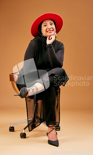 Image of Fashion, fancy and a mature model woman in studio on a brown background sitting on a chair for trendy clothes. Portrait, hat and clothing with an attractive senior female seated to promote style