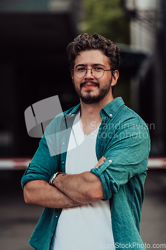 Image of A successful young businessman in a shirt, with crossed arms, poses outdoors, confident expression on his face.