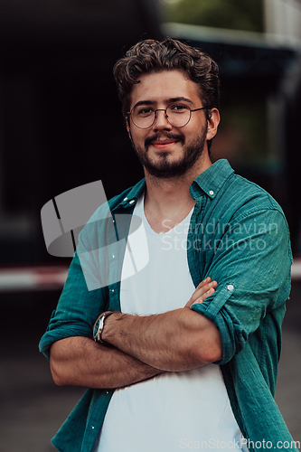 Image of A successful young businessman in a shirt, with crossed arms, poses outdoors, confident expression on his face.