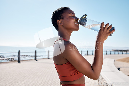 Image of Black woman, drinking water or bottle in fitness workout, training or exercise by beach, ocean or sea in summer location. Smile, happy or sports runner with drink for healthcare wellness recovery