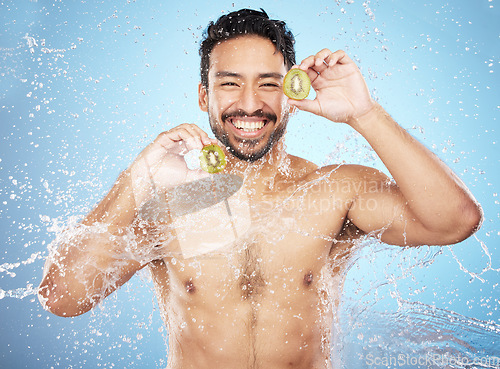 Image of Face portrait, water splash and man with kiwi in studio isolated on a blue background. Cleaning, skincare and hygiene of happy male model washing fresh fruit for vitamin c, nutrition and healthy diet