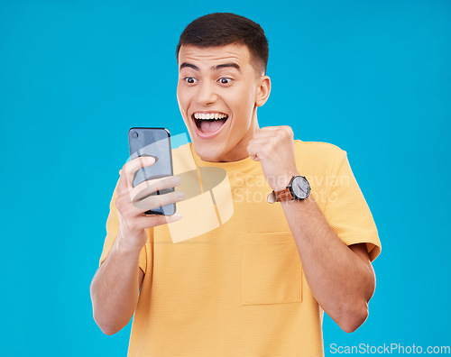 Image of Cellphone, celebration and young man in a studio with fist pump for winning, success or achievement. Happy, smile and male winner from Canada cheering for prize on a phone isolated by blue background