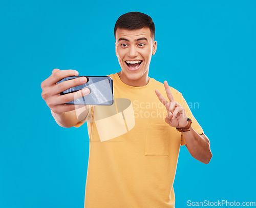 Image of Man, selfie and peace hand sign with happiness, excited with social media with communication on blue background. V emoji, influencer with memory and smile in picture with photography in a studio
