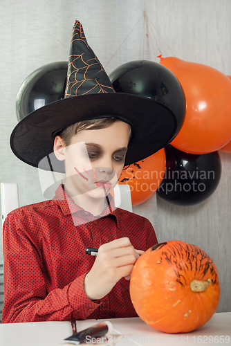 Image of Happy teen boy in costume drawing a pumpkin for the Halloween celebration.