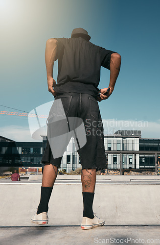 Image of Skate clothes, urban fashion and man with emo, goth or gen z contemporary street style at fitness skate park for training. Blue sky lens flare, concrete building and back view of stylish black person