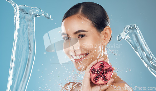 Image of Woman, beauty and water splash with pomegranate, skincare and happy against a blue studio background. Model, smile and shower with fruit for fresh, clean and healthy skin, face or hair with happiness