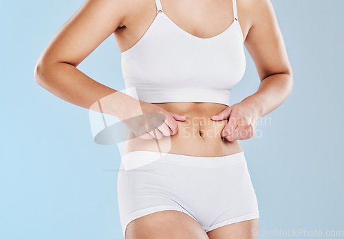 Image of Plus size, fat and woman touching her stomach cellulite in underwear standing in a studio. Overweight, obesity and closeup of a girl model feeling her chubby abdomen isolated by a blue background.