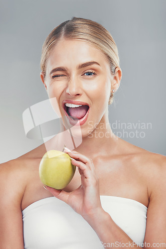 Image of Woman, health and wink portrait with apple for fiber diet, nutrition and wellness lifestyle. Fruit, healthy and digestion dieting marketing with happy model girl on gray studio background.