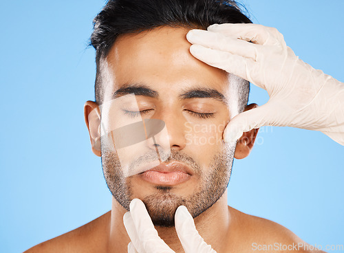 Image of Cosmetic, plastic surgery and face of young man, for skincare and white gloves on blue studio background for dermatology care. Makeup, Indian male and skin rejuvenation for wellness and collagen.