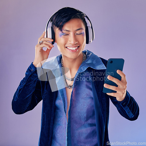 Image of Headphones, studio and gay man with makeup listening to music on the internet with smartphone. Cosmetics, lgbtq and happy guy streaming audio, podcast or radio on phone isolated by purple background.