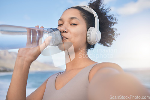 Image of Woman runner, drinking water and selfie with music while outdoor for exercise, workout or fitness. Black woman, headphones and beach for training, wellness or health in nature by ocean in summer sun