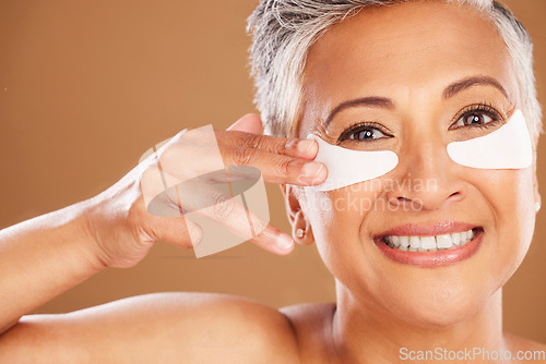 Image of Beauty, skincare and senior woman with eyepads on face for a natural facial routine in studio. Cosmetic, wellness and portrait of elderly lady with skin treatment product isolated by brown background