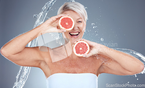 Image of Grapefruit, water and senior woman with a splash for skincare, beauty and wellness against a grey studio background. Water splash, health and excited elderly model with fruit for diet lifestyle