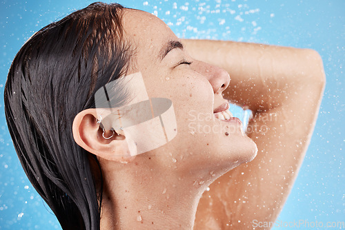 Image of Shower, water and face of a woman cleaning body to relax for wellness against a blue studio background. Hair care, happy and Asian model washing for grooming, hygiene and health with a smile