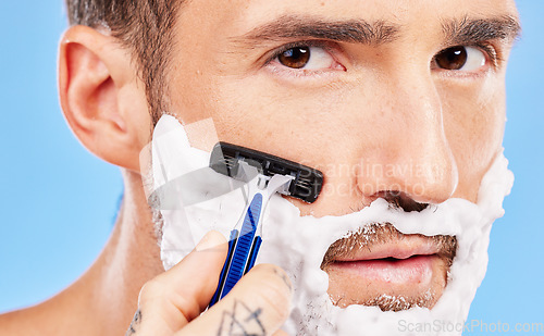 Image of Grooming, cream and man shaving his face with a razor for wellness, skincare and health against a blue studio background. Beauty, young and portrait of a model with foam on beard for hygiene