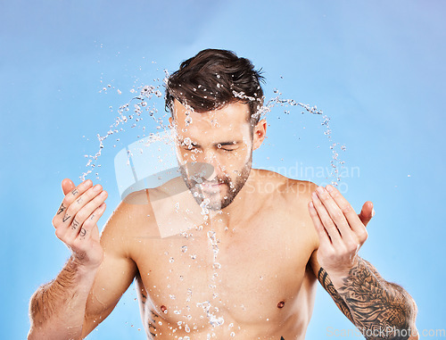 Image of Face, water and splash with a man cleaning his skin in studio on a blue background for hygiene or hydration. Wellness, skincare and water splash with a handsome young male washing his body for care