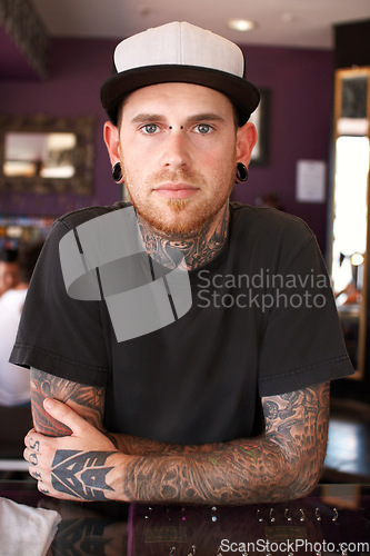 Image of Tattoos, creative and portrait of a man with tattoos standing by the counter of his artistic studio. Confident, cool and punk man with a edgy, funky and body art or ink creativity business store.