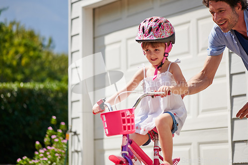 Image of Child learning from dad to ride a bicycle as support, trust and skill development outdoor of a home or house together. Parent, safety and happy father care, help and teaching kid or girl to use bike