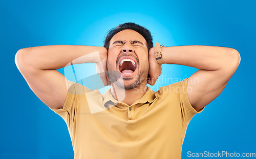 Image of Man cover his ears while shouting in a studio for angry, upset or mad argument expression. Crazy, scream and young male person with an open mouth for loud voice gesture isolated by a blue background.