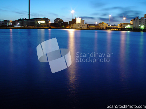 Image of Ocean, night and buildings in port with lights, reflection on calm blue water or holiday location. City at sea in evening, travel, skyline and nature, urban beach village for vacation, peace and view