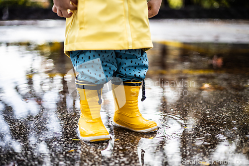 Image of Small infant boy wearing yellow rubber boots and yellow waterproof raincoat standing in puddle on a overcast rainy day. Child in the rain.