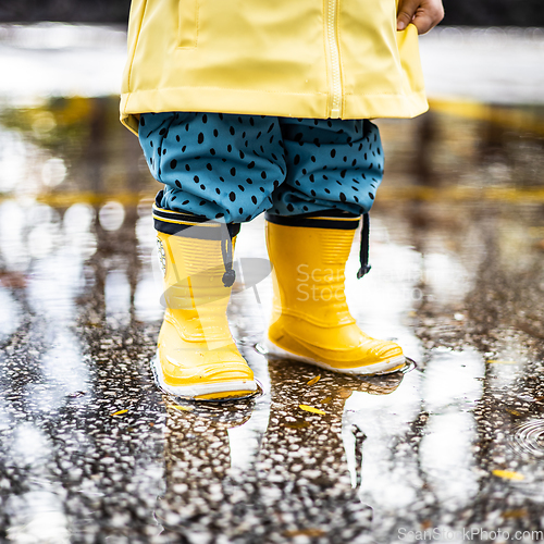 Image of Small infant boy wearing yellow rubber boots and yellow waterproof raincoat standing in puddle on a overcast rainy day. Child in the rain.