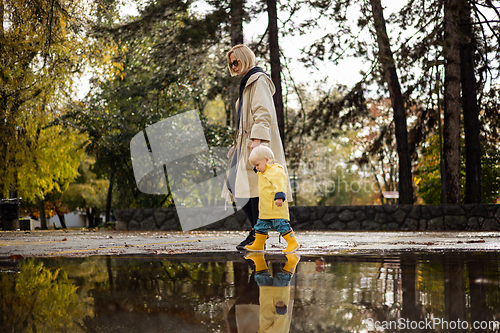 Image of Small bond infant boy wearing yellow rubber boots and yellow waterproof raincoat walking in puddles on a overcast rainy day holding her mother's hand. Mom with small child in rain outdoors.