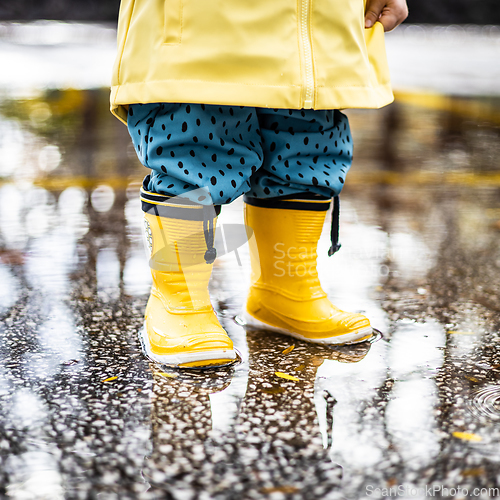 Image of Small infant boy wearing yellow rubber boots and yellow waterproof raincoat standing in puddle on a overcast rainy day. Child in the rain.