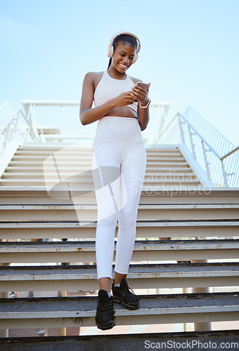 Image of Smile, fitness and black woman on a phone for music, social media content and streaming podcast via headphones. Happy, city and young girl checking progress on a wellness or exercise app in Africa