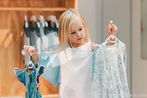 Image of Child, fashion and shopping while choosing kids clothing in a shop, store or boutique with a cute girl holding dresses thinking or picking color choice. Little kid shopper or customer buying clothes