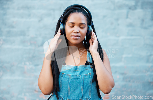 Image of Black woman, headphones and music with a girl feeling calm, relax and content listening to a podcast standing outside against city or urban wall. African female relaxed during mindful audio streaming