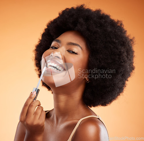 Image of Lipgloss, smile and black woman excited about makeup against an orange studio background. Face portrait of a young African girl model with lipstick and cosmetic beauty product for care of lips