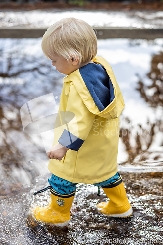 Image of Small bond infant boy wearing yellow rubber boots and yellow waterproof raincoat walking in puddles on a overcast rainy day. Child in the rain.