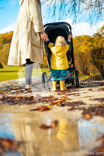 Image of Sun always shines after the rain. Small blond infant boy wearing yellow rubber boots and yellow waterproof raincoat walking in puddles, pushing stroller in city park, holding mother's hand after rain.