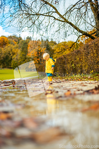 Image of Sun always shines after the rain. Small bond infant boy wearing yellow rubber boots and yellow waterproof raincoat walking in puddles in city park on sunny rainy day.