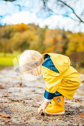 Image of Sun always shines after the rain. Small bond infant boy wearing yellow rubber boots and yellow waterproof raincoat walking in puddles in city park on sunny rainy day.