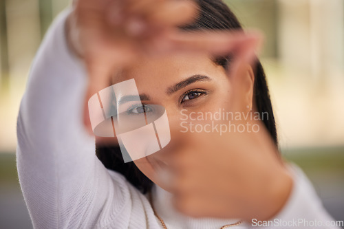 Image of Hands, frame and eyes with a woman looking through her fingers while framing her face outdoor. Portrait, girl and happy with a young female posing behind a hand sign on a blurry background outside