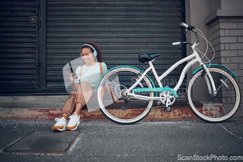 Image of Bike, woman and headphone with phone in city relax, with bicycle and happy texting, social media apps and chat. Girl, lady and with eco friendly transportation, trendy or edgy look sit on sidewalk