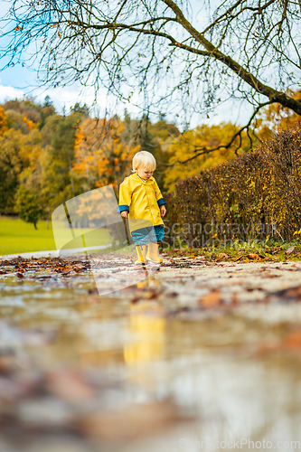 Image of Sun always shines after the rain. Small bond infant boy wearing yellow rubber boots and yellow waterproof raincoat walking in puddles in city park on sunny rainy day.