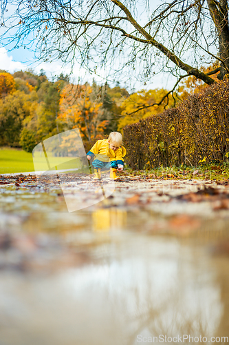 Image of Sun always shines after the rain. Small bond infant boy wearing yellow rubber boots and yellow waterproof raincoat walking in puddles in city park on sunny rainy day.