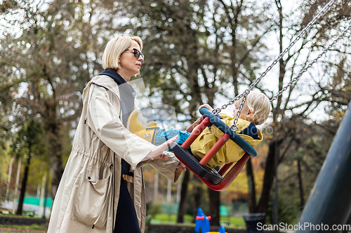 Image of Mother pushing her infant baby boy child wearing yellow rain boots and cape on swing on playground outdoors on cold rainy overcast autumn day in Ljubljana, Slovenia