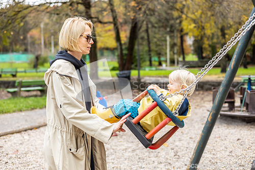 Image of Mother pushing her infant baby boy child wearing yellow rain boots and cape on swing on playground outdoors on cold rainy overcast autumn day in Ljubljana, Slovenia