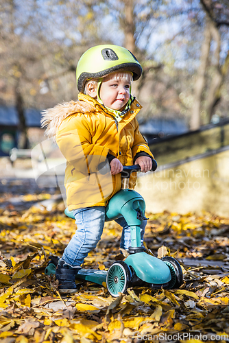 Image of Adorable toddler boy wearing yellow protective helmet riding baby scooter outdoors on autumn day. Kid training balance on mini bike in city park. Fun autumn outdoor activity for small kids.