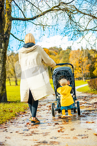 Image of Sun always shines after the rain. Small blond infant boy wearing yellow rubber boots and yellow waterproof raincoat walking in puddles, pushing stroller in city park, holding mother's hand after rain.