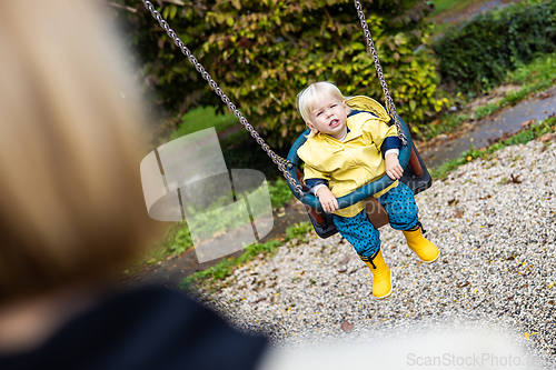 Image of Mother pushing her infant baby boy child wearing yellow rain boots and cape on swing on playground outdoors on cold rainy overcast autumn day in Ljubljana, Slovenia