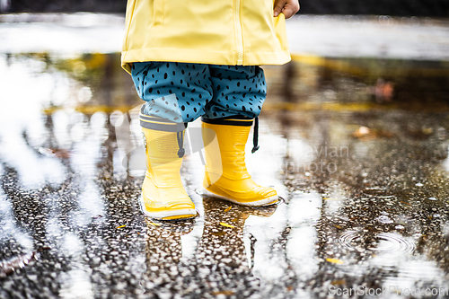 Image of Small infant boy wearing yellow rubber boots and yellow waterproof raincoat standing in puddle on a overcast rainy day. Child in the rain.