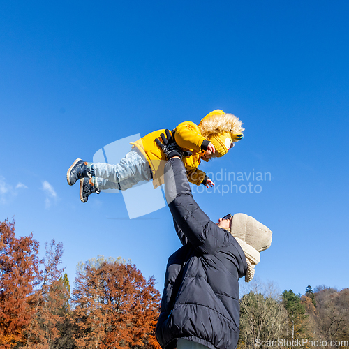 Image of More, more,...mum, that's fun. Happy young mother throws her cute little baby boy up in the air. Mother's Day, Mather and her son baby boy playing and hugging outdoors in nature in fall.