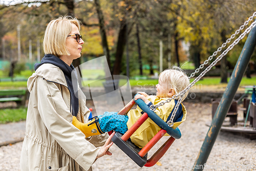 Image of Mother pushing her infant baby boy child wearing yellow rain boots and cape on swing on playground outdoors on cold rainy overcast autumn day in Ljubljana, Slovenia