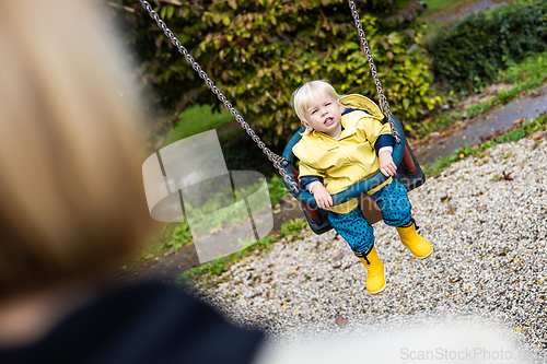 Image of Mother pushing her infant baby boy child wearing yellow rain boots and cape on swing on playground outdoors on cold rainy overcast autumn day in Ljubljana, Slovenia